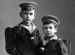 Two young boys in complete sailor suits and officers hats pose for formal portrait.