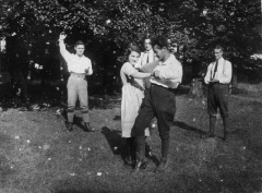 A man and a woman folk dance on a lawn as three other men clap along.