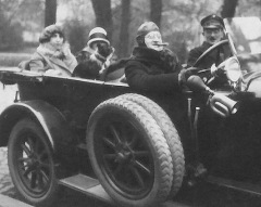 A cigar chomping man wearing a pilot helmet drives an early 19th Century phaeton with its top down as his chauffeur sits beside him and two ladies occupy the rear seat holding a small bulldog