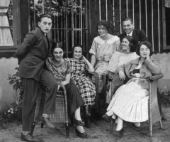 Group portrait out of doors: five ladies in festive dresses seated, two young gentlemen in suit and tie standing.