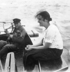 Lady dressed casually seat on gunwale of small boat piloted by man handling outboard motor while cradling a dog.