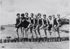 Ten young ladies in mid-1920s bathing attire lined up  each kicking ableft leg high for the  lady in front to grab onto.