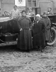 Six well-bundled-up in long coats and wearing close-fitting hats, pose in front of the open-top 1920s black automobile.