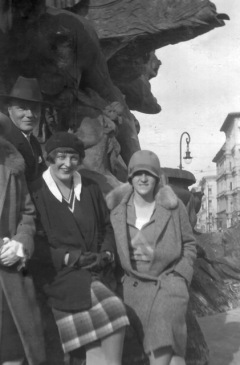 Two ladies in warm fashionable coats and hats pose against a large statue, Berlin street scene behind.