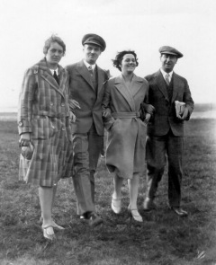 Two young women arm-in-arm with two young men strolling open field.