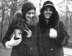 Two young ladies laughing and hugging dressed in furs and hats in chilly part, one holding a small box movie camera.