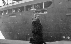 Woman in long coat poses beside dockside ship.