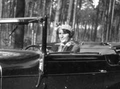 Stylishly dressed lady at the wheel of her 1930s convertible near forested area.