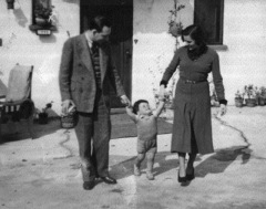 1930s couple in front of small house, each grasping a hand of their toddler boy who looks up to them.