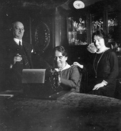 Seated woman operates a typewriter as a younger woman and older man stand behind her looking on.