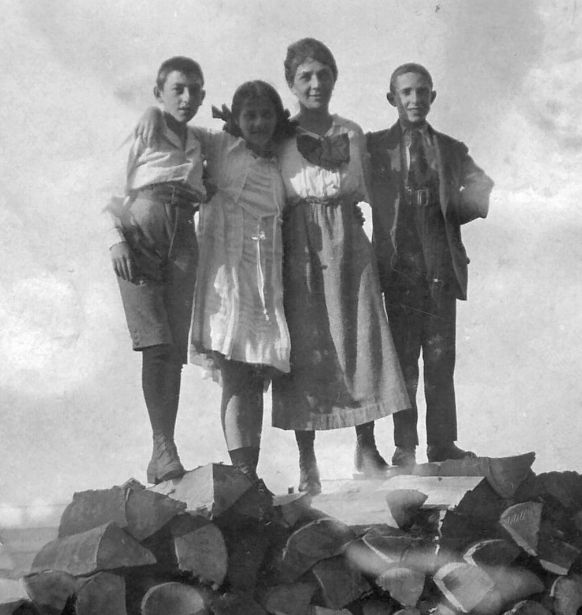 A woman and three young children stand side-by-side atop a pile of split firewood arms around each other.