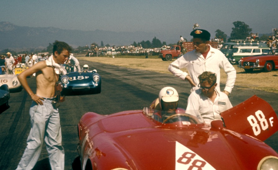 1958 photo of sleek race cars lined up for a race in Santa Barbara Airport featuring a youthful Tim Constantine approaching driver Jack McAfee in a red 550 Porsche Spyder and McAfee's mechanic, Vasek Polak. 