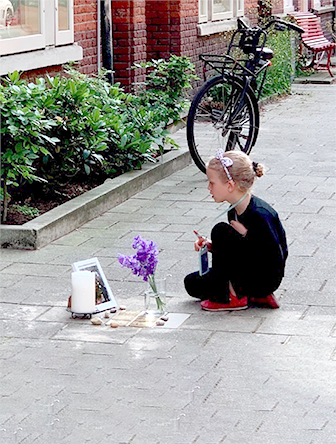 Young girl sits gazing at memorial arrangement of a book, a candle, several small stones, and flowers in a vase arranged around sidewalk-mounted Stolperstein, or "stepping stones".