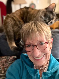 Smiling leslie zurla in her home office with her large sleepy cat looking over her shoulder.